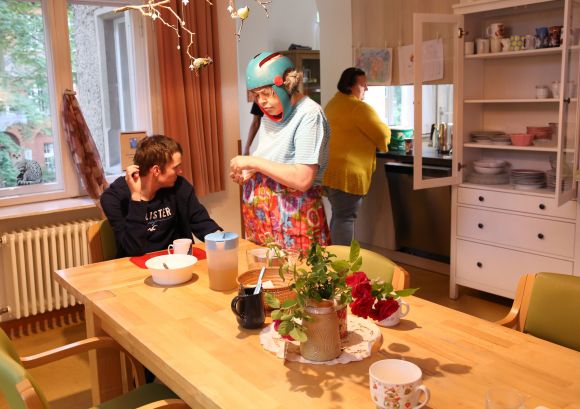 Wohnsituation in der Küche: eine Mitbewohnerin deckt den Tisch, eine andere spült ab, ein junger Mann istzt am gedeckten Tisch