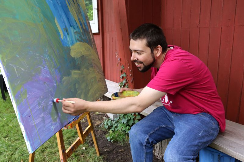 Ein junger Mann sitzt auf einer Bank vor seiner Leinwand und malt lächelnd ein Gemälde