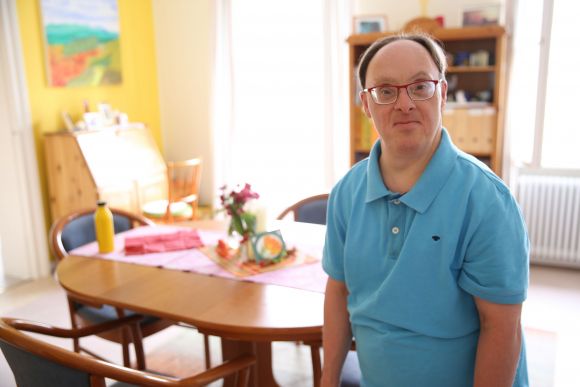 Ein Bewohner unserer LebensOrte im blauen Polo-Shirt steht in seinem Wohnzimmer und schaut lächelnd in die Kamera